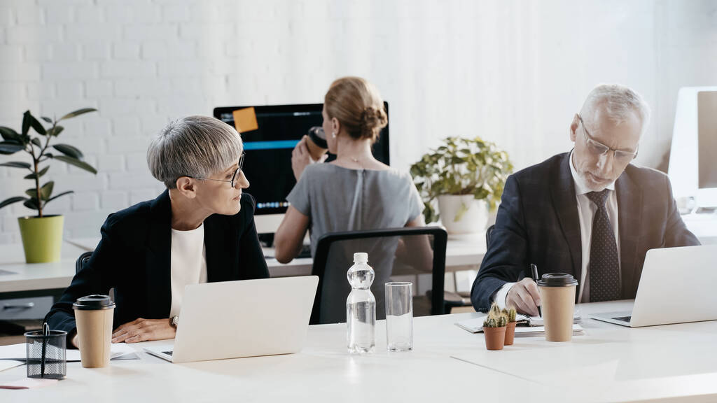 Бізнесмен у формальному носінні пише на ноутбуці біля пристроїв та колег в офісі
  - Фото, зображення