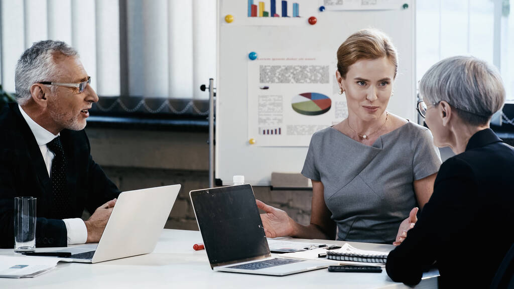 Бізнес-леді дивиться на зрілого колегу біля пристроїв під час зустрічі в офісі
  - Фото, зображення