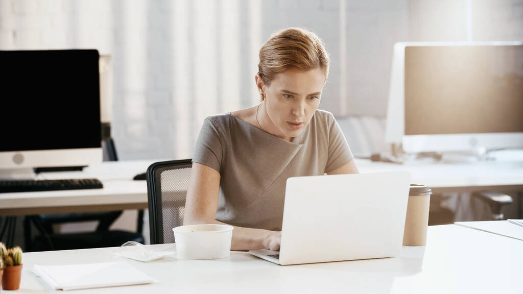 Bizneswoman za pomocą laptopa w pobliżu na wynos kawa i pojemnik w biurze  - Zdjęcie, obraz