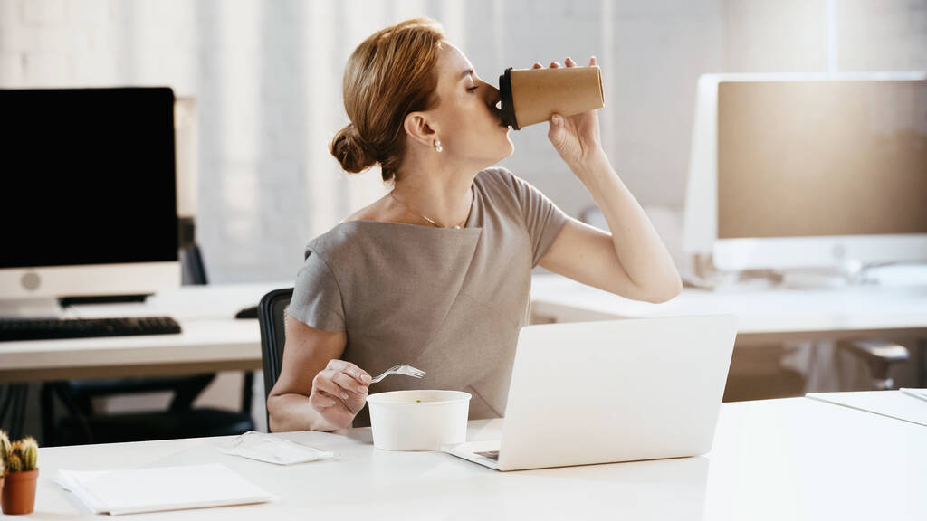 Вид сбоку на предпринимательницу, пьющую кофе, чтобы подойти к свежему салату и ноутбуку в офисе  - Фото, изображение