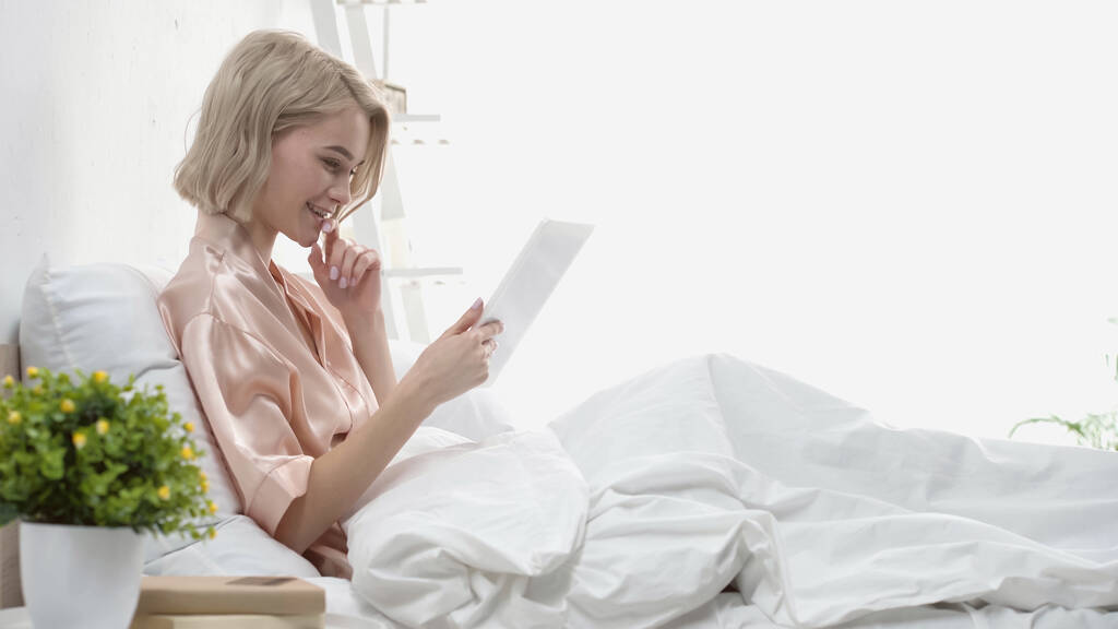 デジタルタブレットで幸せなブロンドの女性を眺めベッドで笑顔で  - 写真・画像