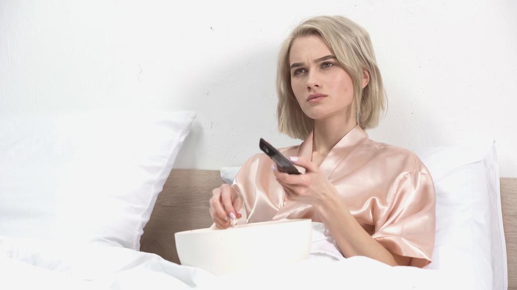 νεαρή ξανθιά γυναίκα κάθεται με μπολ κρατώντας τηλεχειριστήριο και βλέποντας ταινία στο κρεβάτι  - Φωτογραφία, εικόνα