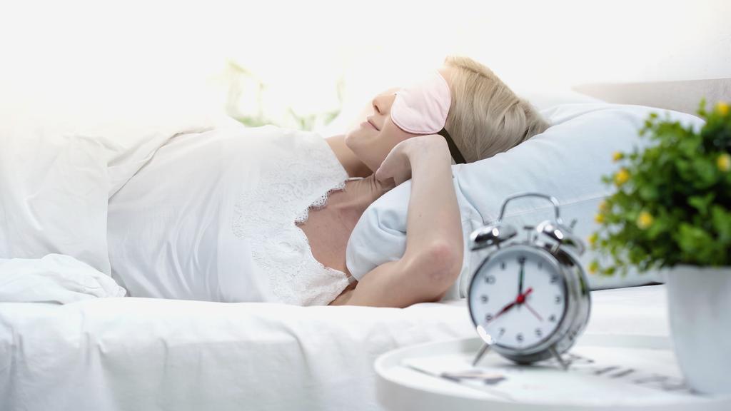 junge blonde Frau mit Schlafmaske im Bett liegend, streckend und lächelnd im Schlafzimmer - Foto, Bild