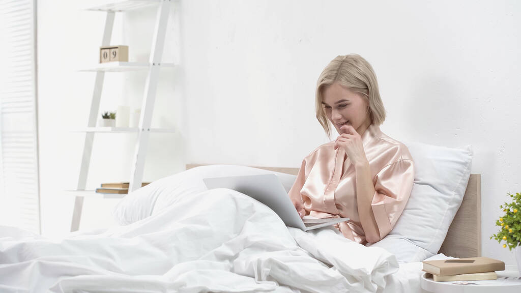 glimlachende blonde freelancer met behulp van laptop terwijl zitten in bed - Foto, afbeelding