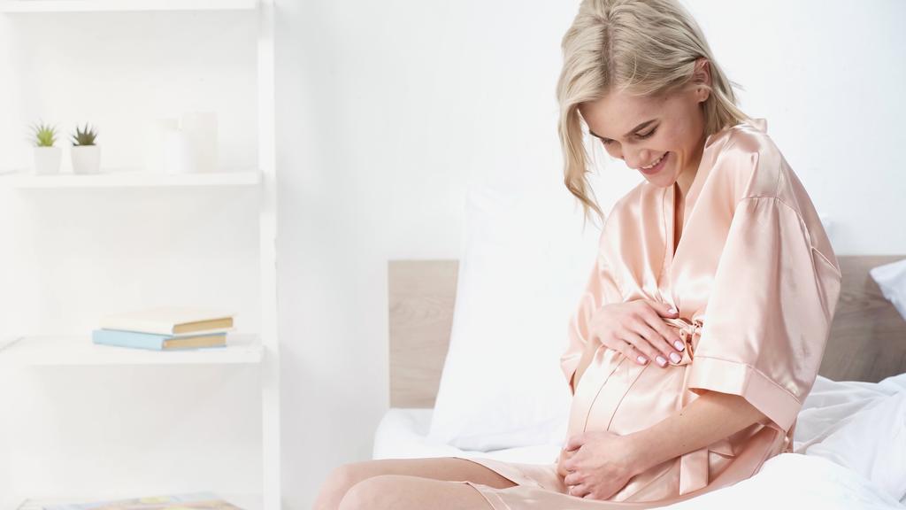 fröhliche Schwangere berührt Bauch und lächelt im Schlafzimmer - Foto, Bild