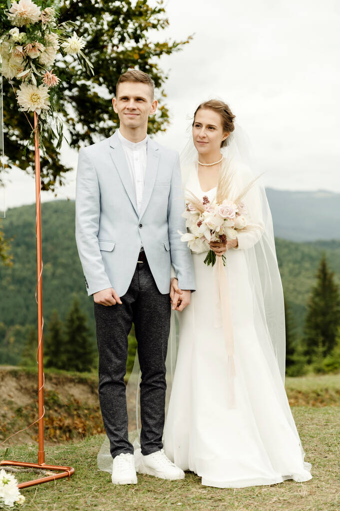 νύφη και γαμπρός κοντά στην αψίδα κατά τη διάρκεια της τελετής του γάμου. γαμήλιο ζευγάρι στα βουνά κοντά σε ένα κλαδί δέντρου - Φωτογραφία, εικόνα