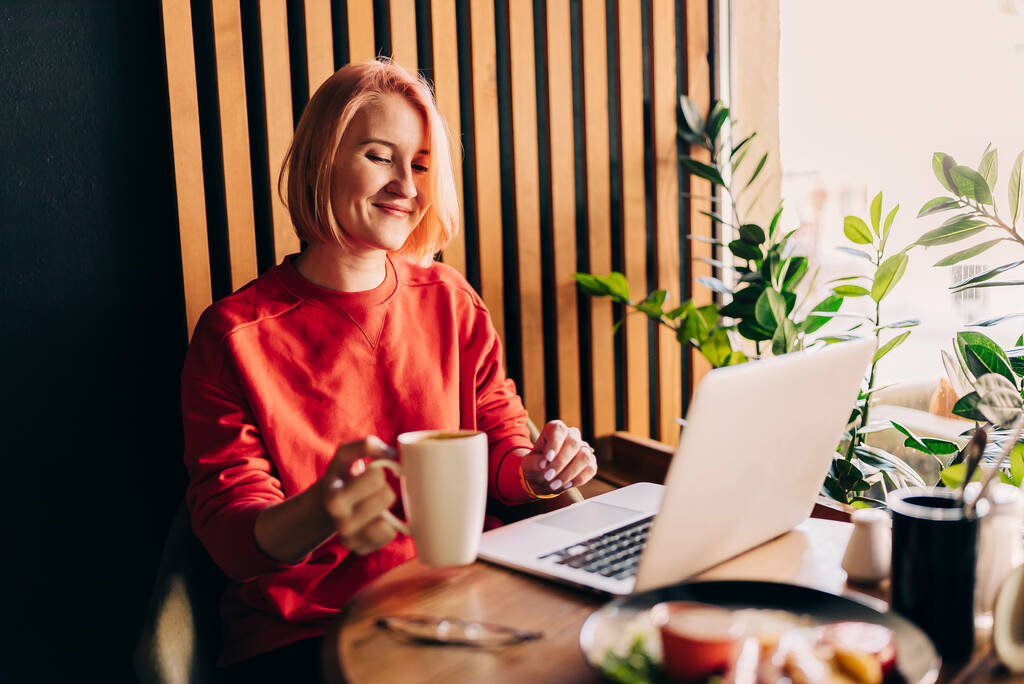 Nuori blondi nainen yllään punainen rento villapaita kahvilassa työskentelee kannettavan tietokoneen ja juo kahvia - Valokuva, kuva