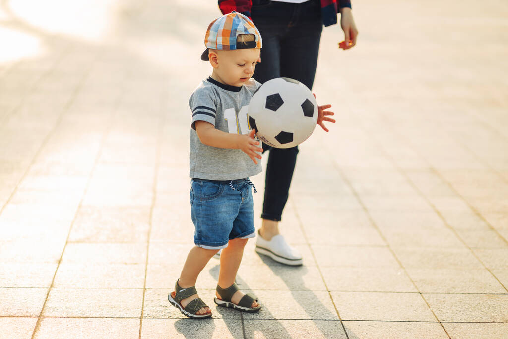 Мама и сын играют в футбол на открытом воздухе в парке, маленький мальчик пинает футбольный мяч, ребенок учится играть в футбол. Семья играет в футбол вместе в парке - Фото, изображение