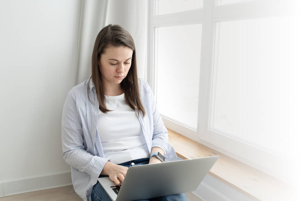 Μεσήλικη γυναίκα που εργάζεται από το σπίτι. Κορίτσι που χρησιμοποιεί φορητό υπολογιστή στο σπίτι, κάθεται κοντά σε ένα μεγάλο παράθυρο. Έννοια απομακρυσμένης εργασίας - Φωτογραφία, εικόνα