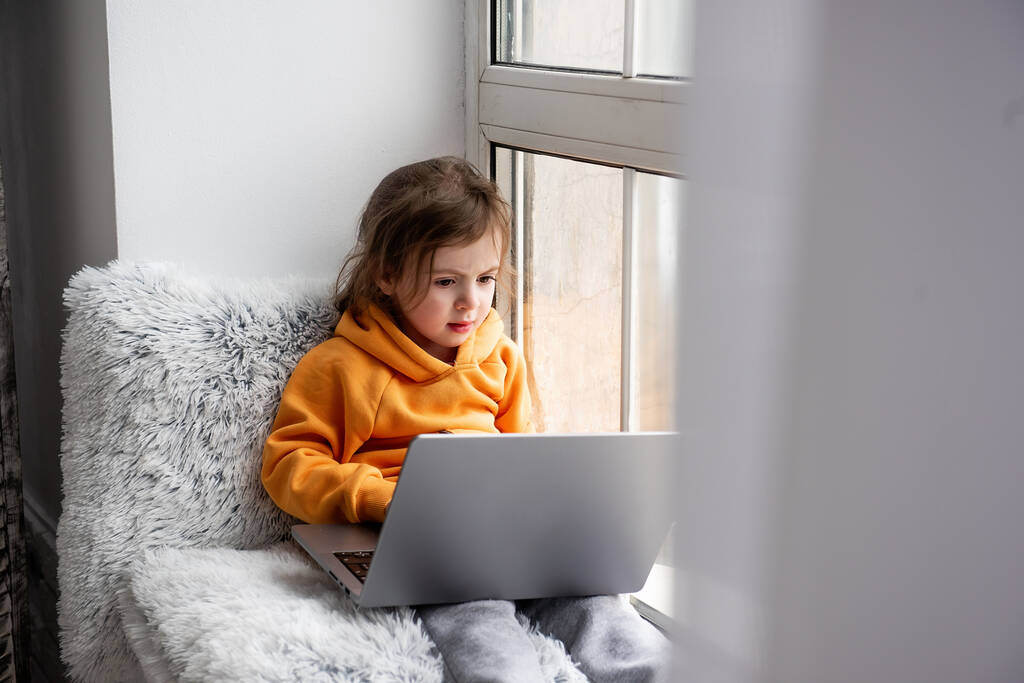 Little toddler girl sits on wide windowsill, plays games on laptop, listens to music on headphones. Child in an orange hoodie, gray pants at the window of the house. Distance learning for preschoolers - Photo, Image
