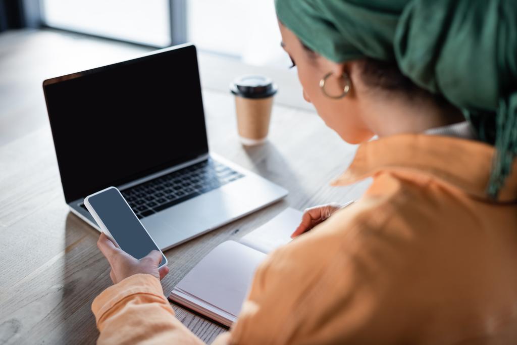 Bulanık Müslüman kadın elinde bilgisayar ve dizüstü bilgisayarın yanında boş ekranlı akıllı telefon tutuyor. - Fotoğraf, Görsel