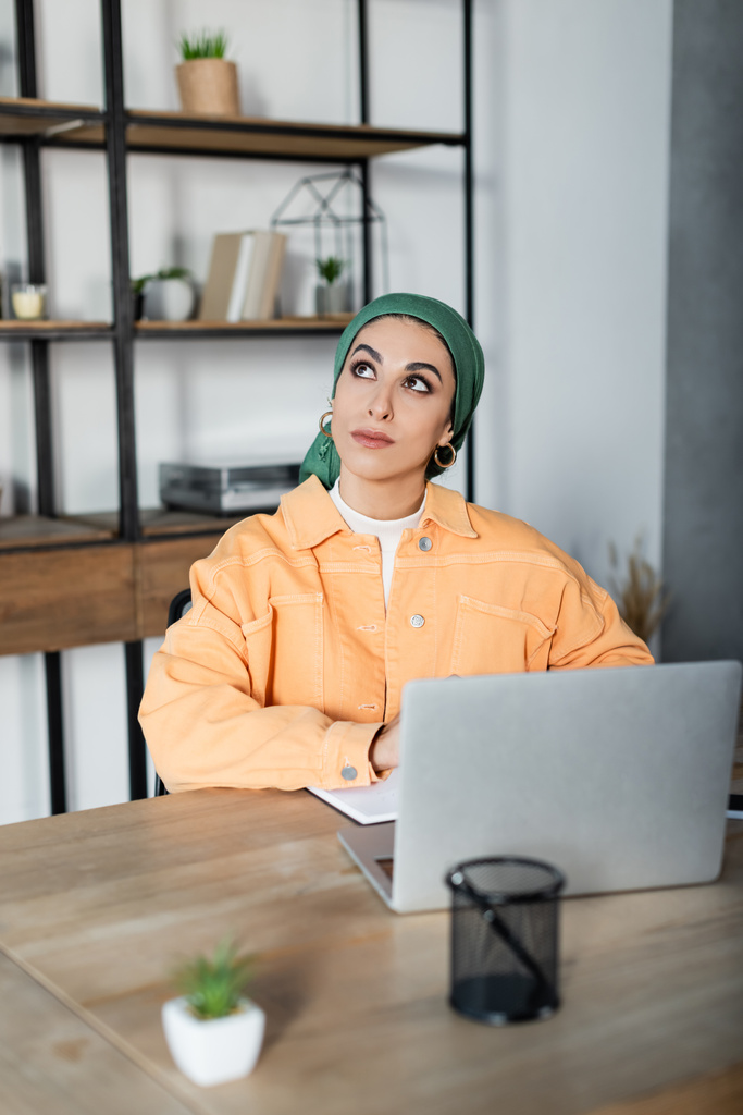 dreamy muslim woman looking away while sitting near laptop at home - Photo, Image