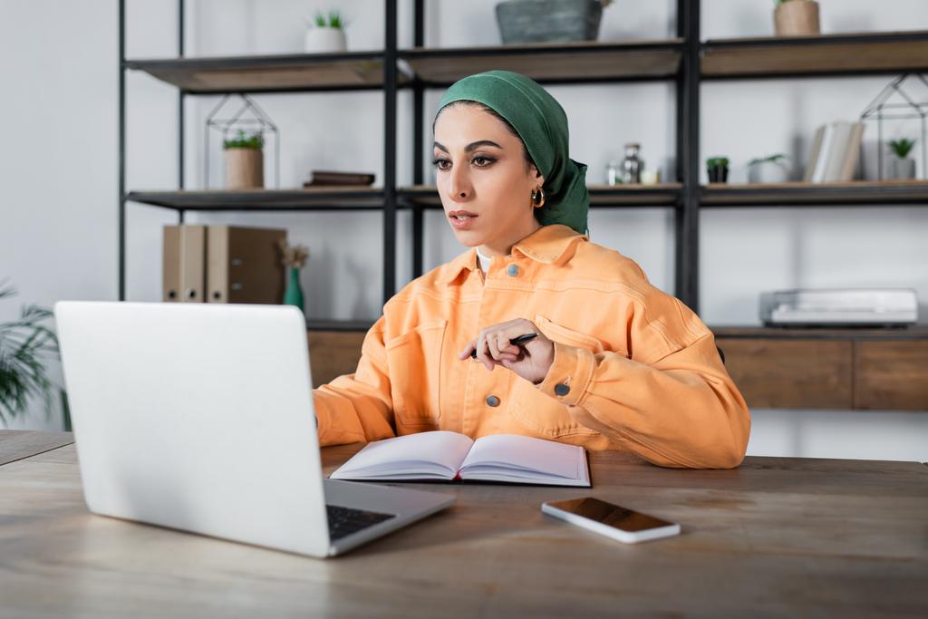 arabialainen nainen otsikossa katsomassa kannettavaa tietokonetta lähellä tyhjää muistikirjaa pöydällä - Valokuva, kuva