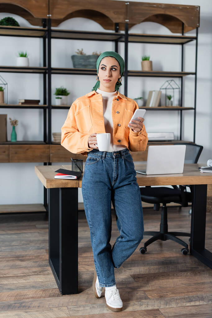 vista completa de la mujer musulmana con taza de té y teléfono inteligente de pie cerca del escritorio en casa - Foto, imagen