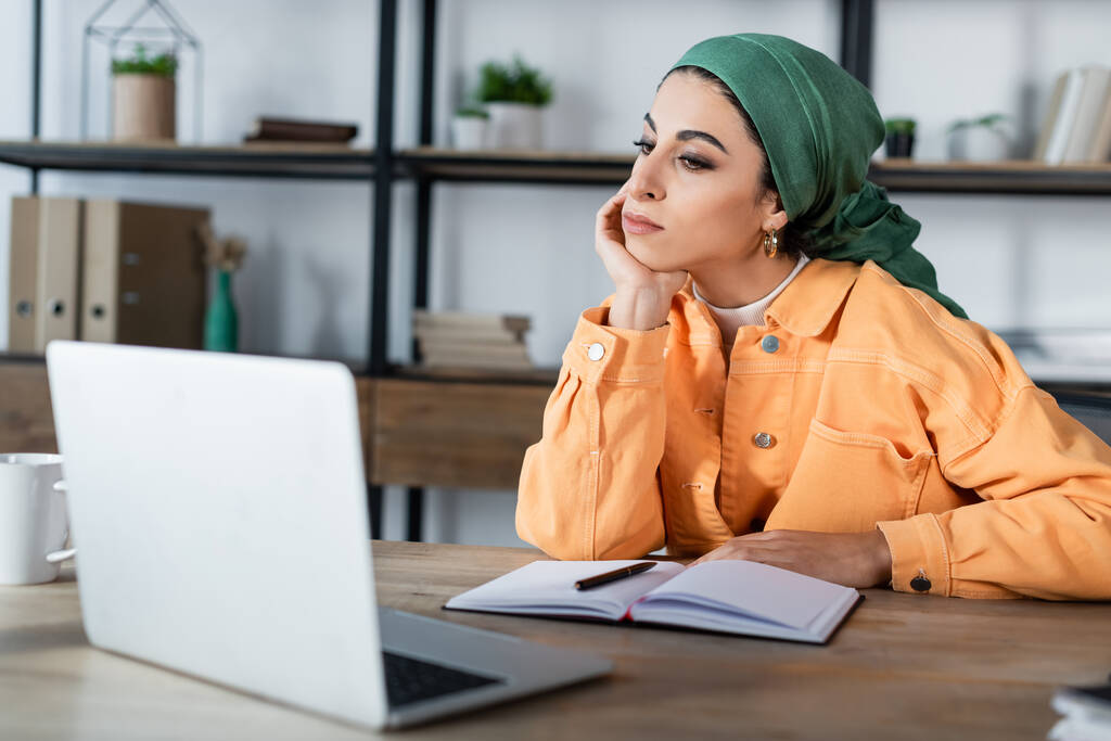 muslim γυναίκα κάθεται με το χέρι κοντά στο πρόσωπο κατά τη διάρκεια του online μαθήματος στο φορητό υπολογιστή στο σπίτι - Φωτογραφία, εικόνα