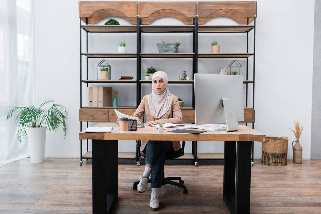 mujer musulmana mirando a la cámara mientras está sentado cerca de la computadora y los documentos en la oficina en casa - Foto, imagen