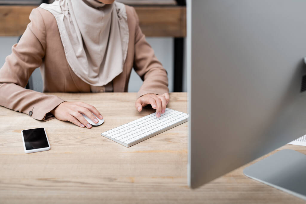 обрізаний вид жінки, що працює на комп'ютері біля смартфона з порожнім екраном на столі
 - Фото, зображення