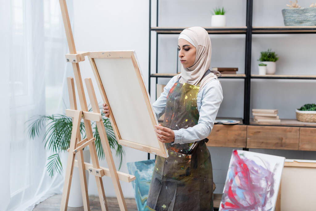 muzułmanka w hidżabie i fartuchu trzymająca płótno w pobliżu sztalugi w domu - Zdjęcie, obraz