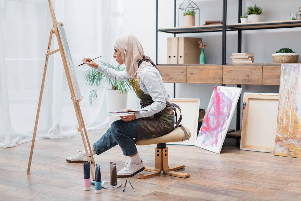 vista lateral de la mujer musulmana sentada en la silla y dibujo en caballete en casa - Foto, Imagen