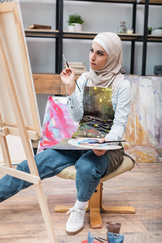 mujer árabe con pincel y paleta mirando lienzo sobre caballete - Foto, Imagen