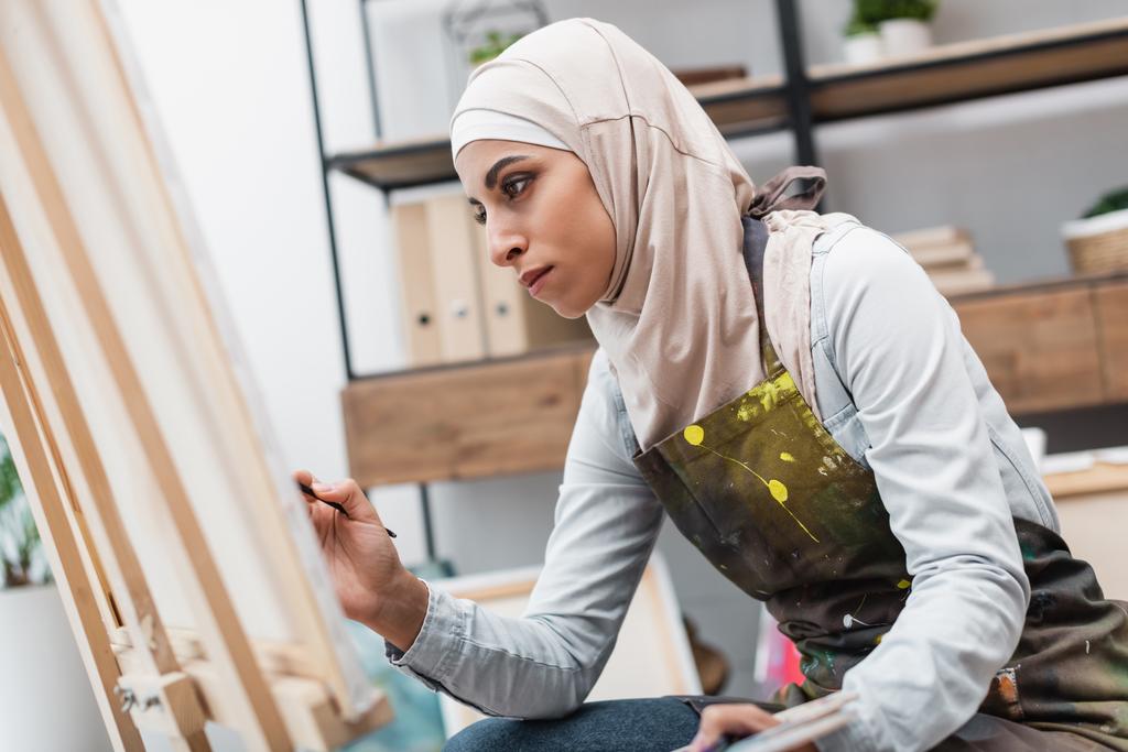 young arabian woman in hijab painting on blurred canvas at home - Photo, Image