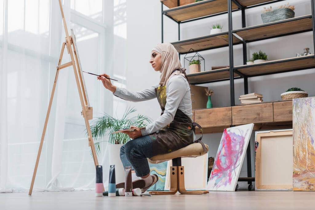 πλαϊνή άποψη των μουσουλμάνων γυναίκα σχέδιο στο καβαλέτο στο στούντιο στο σπίτι - Φωτογραφία, εικόνα
