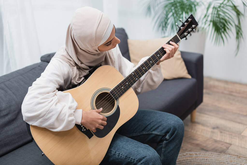 jeune femme musulmane assise sur le canapé à la maison et jouant de la guitare - Photo, image