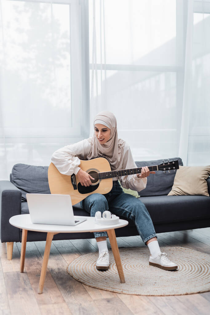 Lächelnde muslimische Frau, die auf der Couch sitzt und neben dem Laptop Gitarre spielen lernt - Foto, Bild