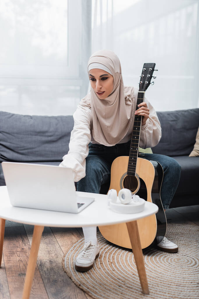 giovane donna musulmana seduta vicino al computer portatile e con in mano la chitarra acustica  - Foto, immagini