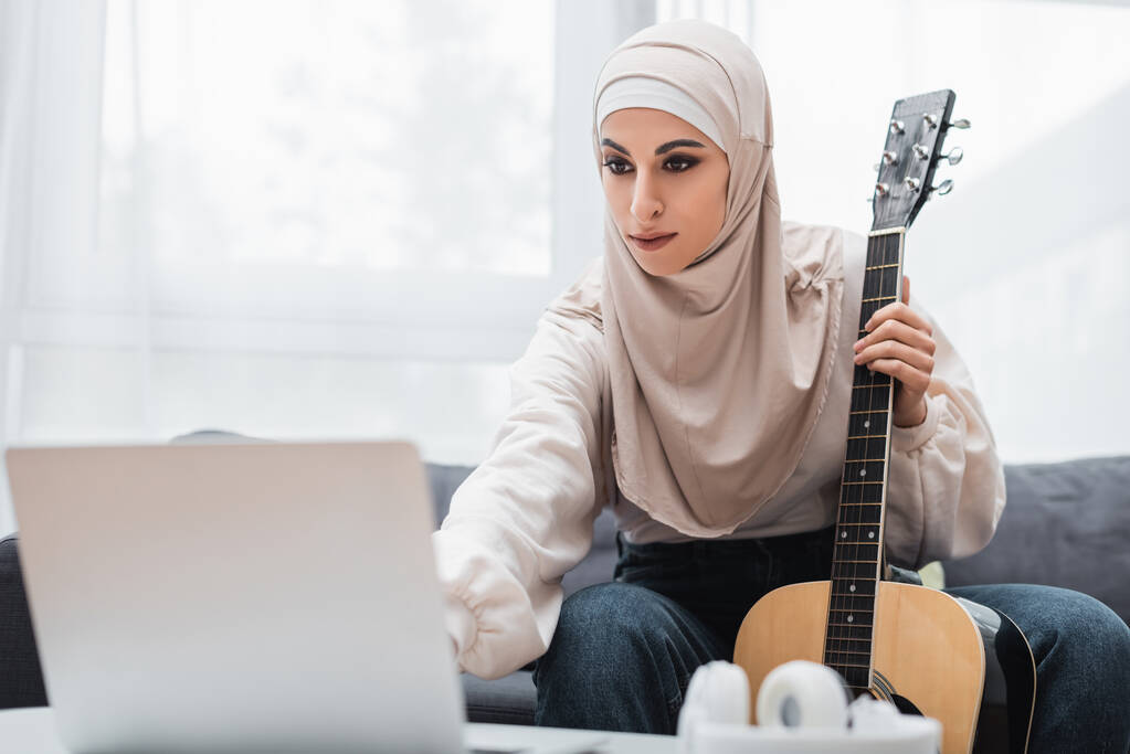 arabian woman in hijab holding guitar and looking at blurred laptop at home - Photo, Image