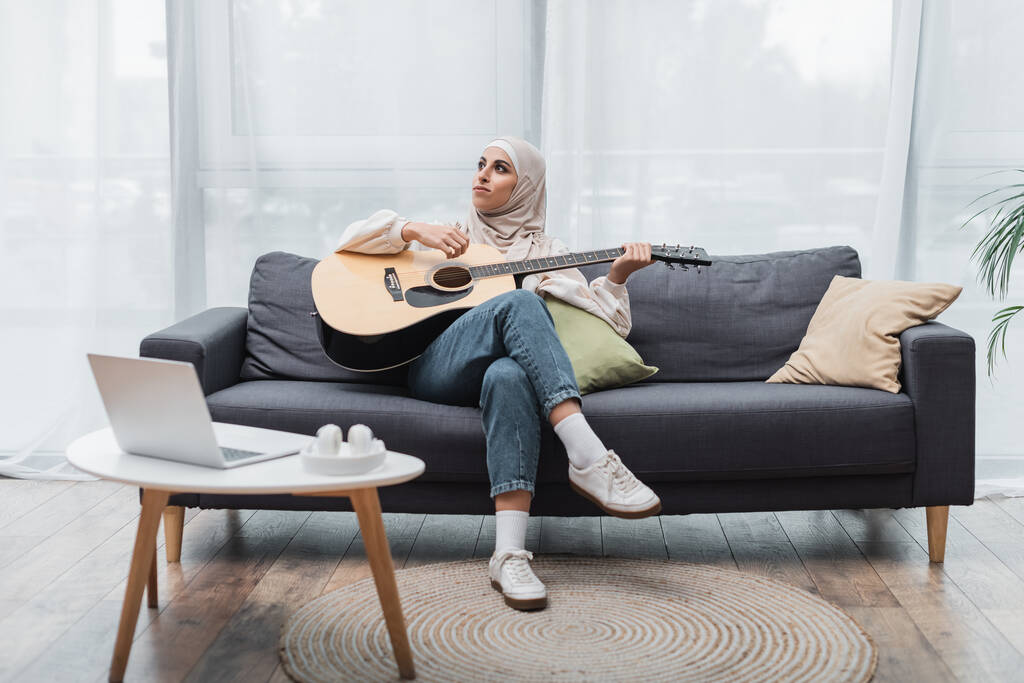 visão de comprimento total da mulher muçulmana olhando para longe enquanto tocava guitarra no sofá perto do laptop - Foto, Imagem