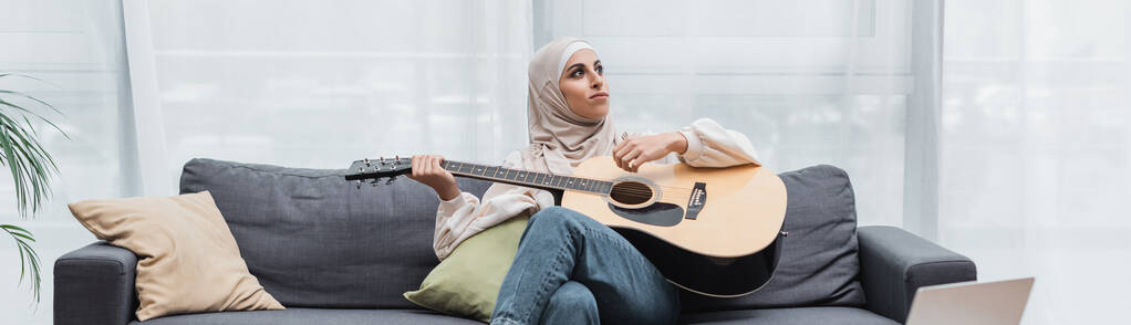 mulher árabe olhando para longe enquanto sentado no sofá e tocando guitarra, banner - Foto, Imagem
