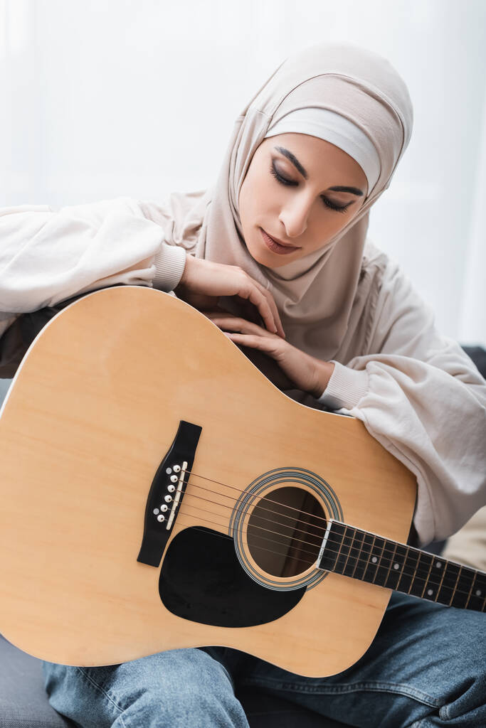 ヒジャーブの若いアラビア人女性が家に座っている間にアコースティックギターにもたれて - 写真・画像
