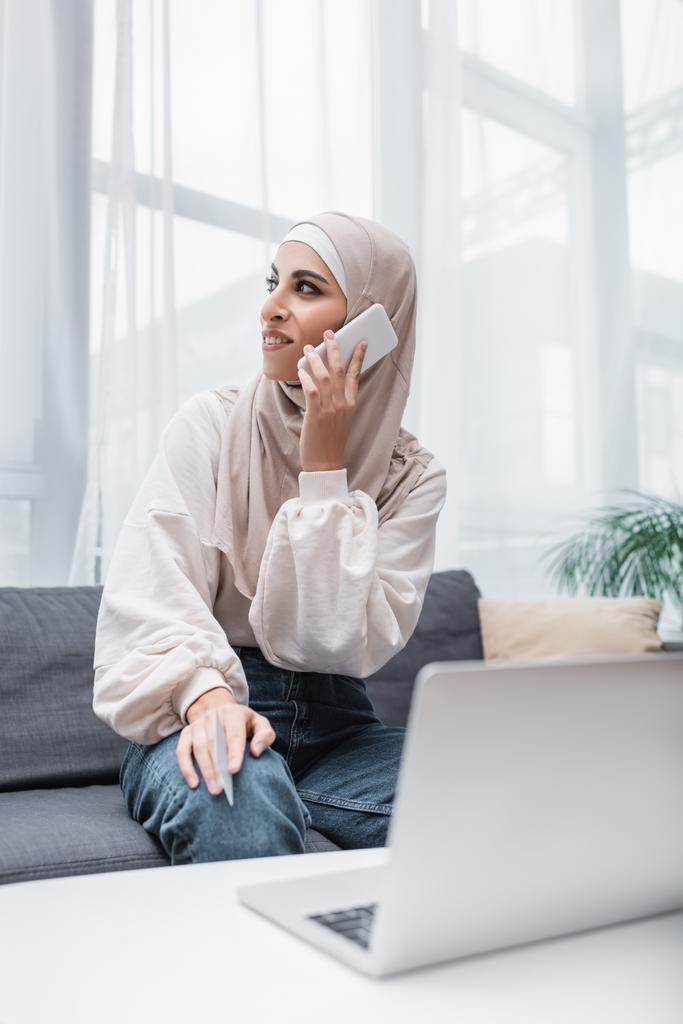 smiling arabian woman with credit card making order on smartphone while sitting near laptop - Photo, Image