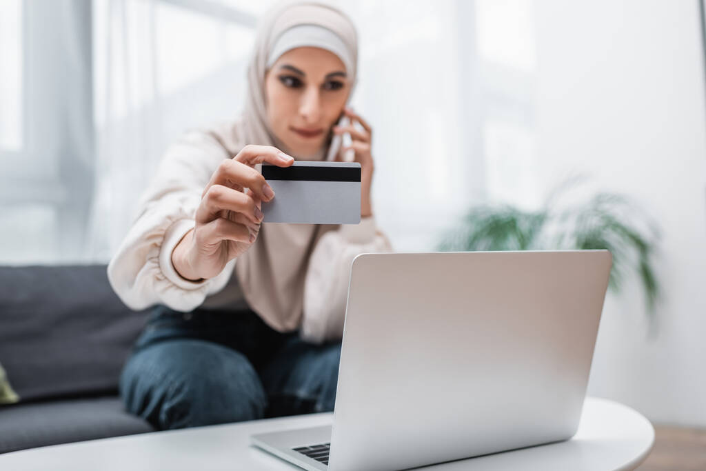 избирательный фокус кредитной карты в руке размытой арабской женщины, звонящей на смартфон рядом с ноутбуком - Фото, изображение