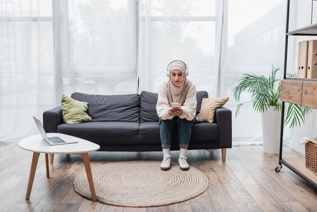 mujer musulmana en auriculares con teléfono inteligente mientras escucha música en la sala de estar - Foto, imagen