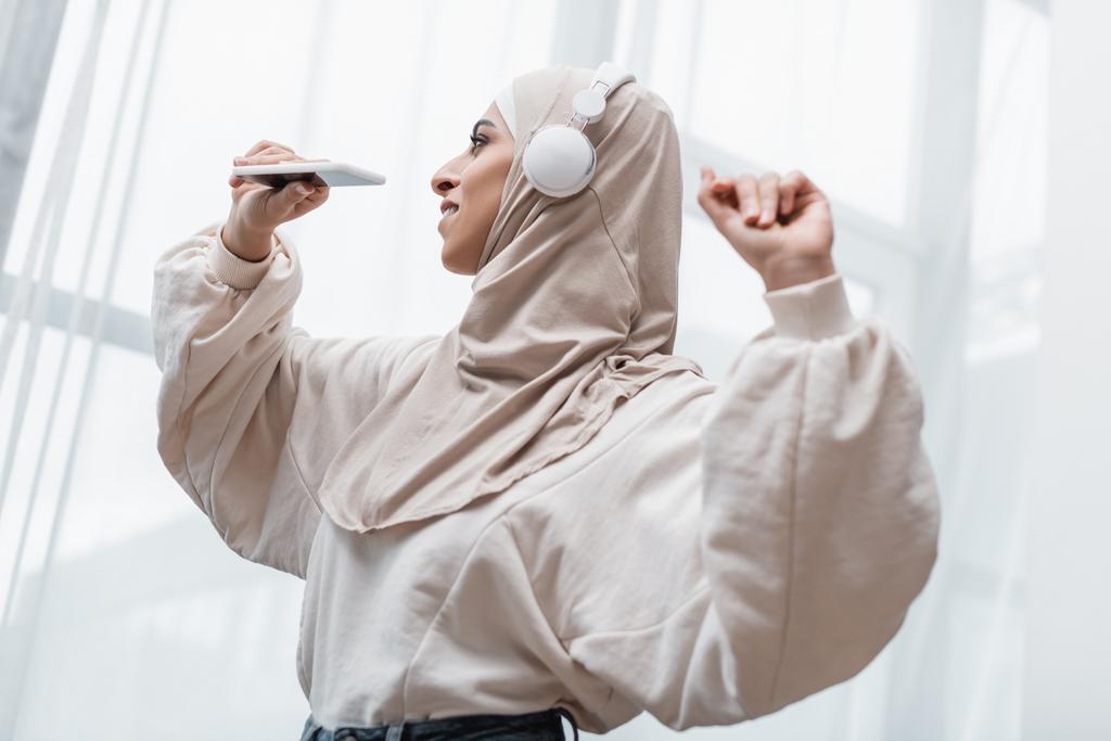 onnellinen arabialainen nainen hijabissa ja kuulokkeet tilalla älypuhelin tanssiessaan kotona - Valokuva, kuva