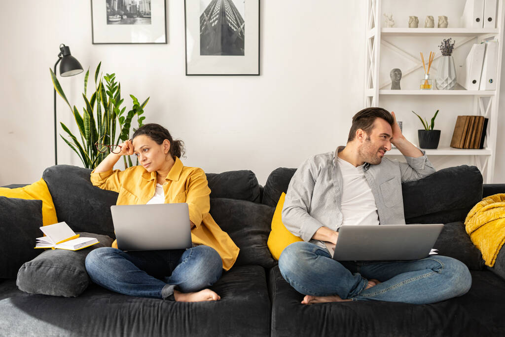 Zmęczona pracą na odległość para w średnim wieku siedząca osobno na kanapie z laptopami - Zdjęcie, obraz