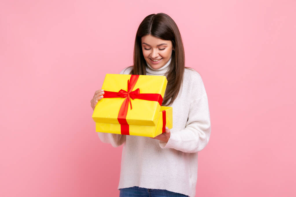 Счастливая темноволосая девушка, выглядывающая в обернутой коробке, открытый подарок, выражающая позитив, в белом свитере в стиле casual. Крытая студия снята на розовом фоне. - Фото, изображение
