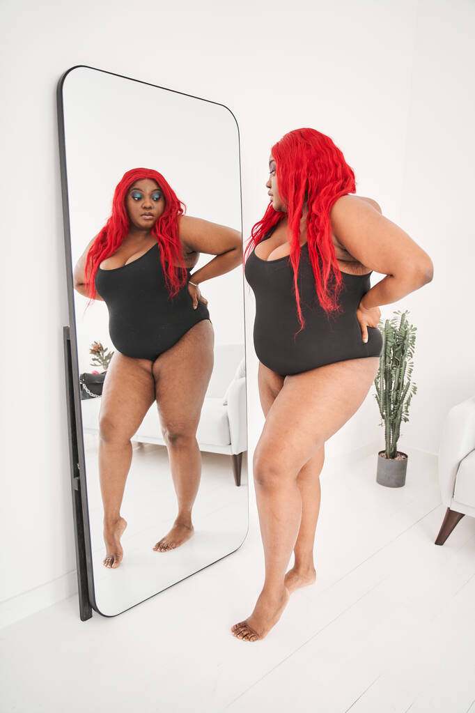 Krásné křivky nadrozměrné multiraciální žena stojí před zrcadlem a dívá se na odraz - Fotografie, Obrázek