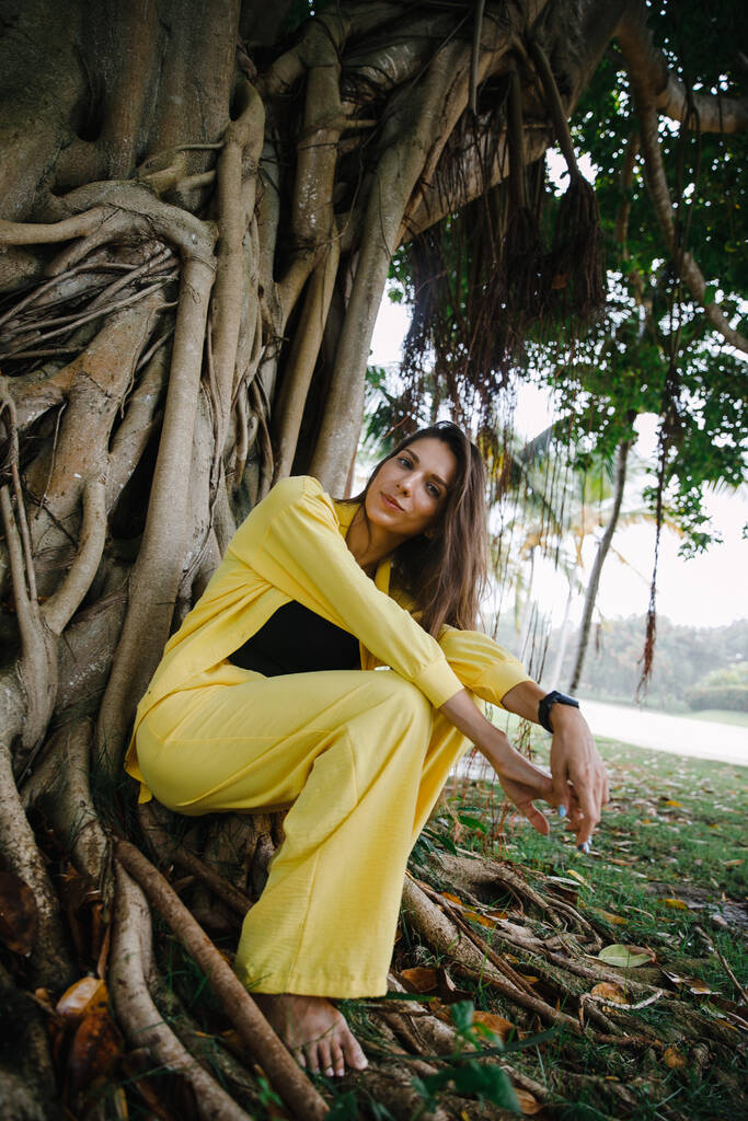 ένα λεπτό μελαχρινό κορίτσι με κίτρινο κοστούμι ποζάρει κομψά σε ένα μπαστούνι του γκολφ σε ένα τροπικό νησί κοντά σε ένα τεράστιο δέντρο στη Δημοκρατία της Δομινικανής - Φωτογραφία, εικόνα