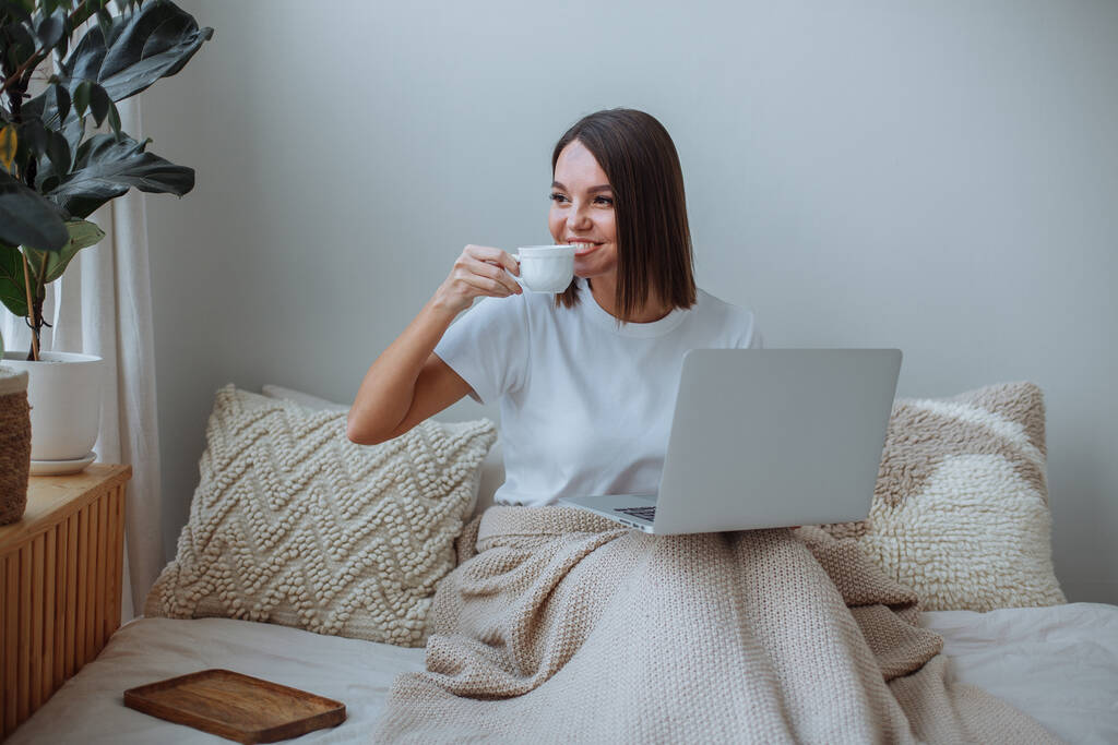 Χαμογελαστή νεαρή μελαχρινή γυναίκα με κοντό κούρεμα σε λευκό t-shirt και μπεζ παντελόνι κάθεται στο κρεβάτι στο σπίτι και δουλεύει σε φορητό υπολογιστή. Πίνω καφέ από λευκή κούπα - Φωτογραφία, εικόνα