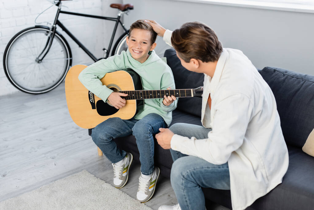 Hombre tocando sonriente hijo con guitarra acústica en casa  - Foto, Imagen