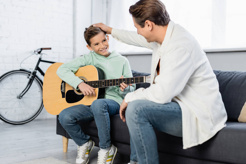 Uśmiechnięty ojciec dotykający włosów syna z gitarą akustyczną na kanapie  - Zdjęcie, obraz