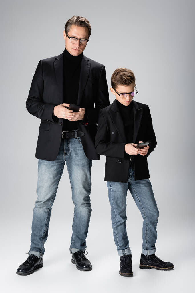 灰色の背景にスマートフォンを使用して人間と十代の息子の完全な長さ  - 写真・画像