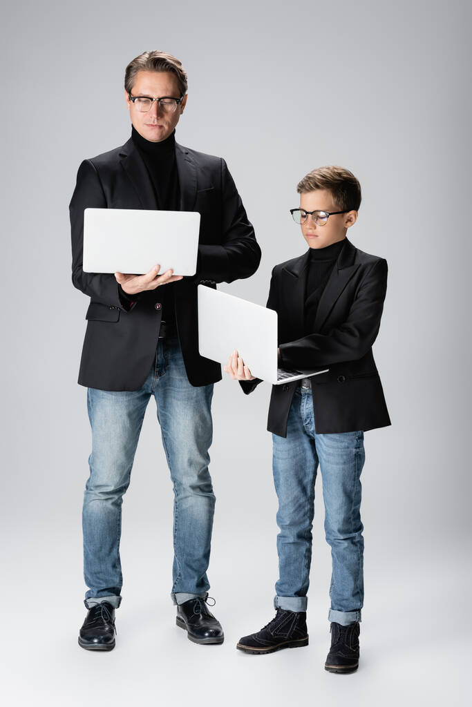 Πλήρες μήκος του trendy άνδρα και αγόρι χρησιμοποιώντας φορητούς υπολογιστές σε γκρι φόντο  - Φωτογραφία, εικόνα