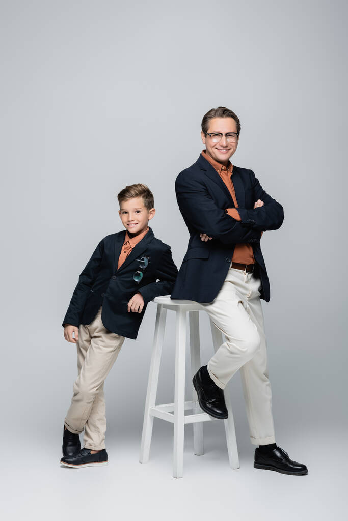 Positiv und stilvoll posieren Vater und Sohn in der Nähe von Stuhl auf grauem Hintergrund  - Foto, Bild