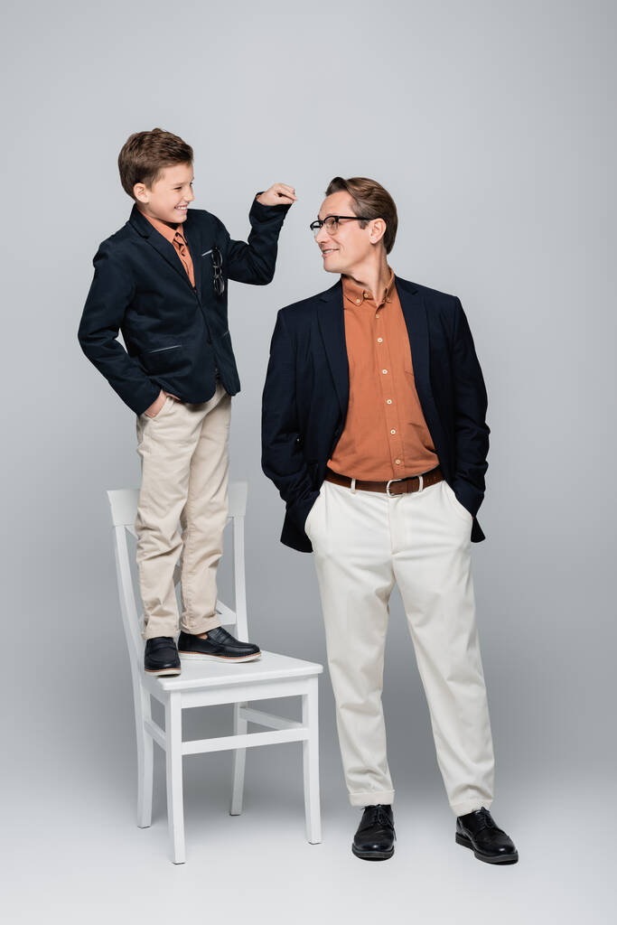 Smiling boy pointing at stylish dad while standing on chair on grey background  - Photo, Image