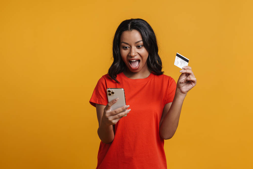 Negro excitado mujer gritando mientras sostiene la tarjeta de crédito y el teléfono celular aislado sobre fondo amarillo - Foto, Imagen