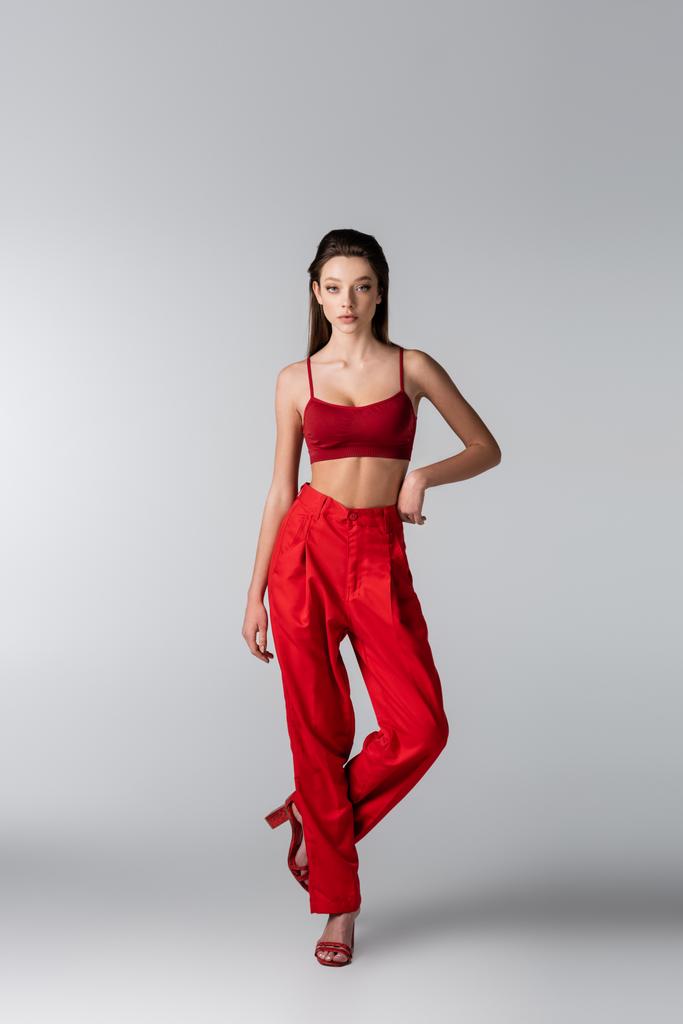 comprimento total do modelo jovem em roupa vermelha posando com a mão no quadril no cinza - Foto, Imagem
